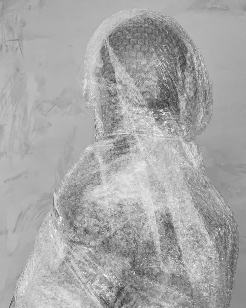 A human mannequin completely wrapped in layers of bubble wrap. The figure stands in profile, facing slightly away from the camera. Monochromatic photograph