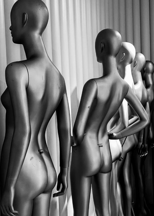 A row of naked mannequins from behind, all with feminine body shapes, in a static position, side by side. 
              They are arranged in a diagonal line extending from the foreground to the background of the image.Monochromatic photograph